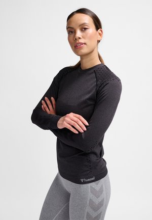 Frau mit verschränkten Armen, die einen dunklen Langarmtop und hellgraue Leggings mit Chevron-Mustern trägt, stehend vor einem einfarbigen weißen Hintergrund.