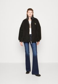 Giacca imbottita nera con colletto alto, zip dorata e dettagli con emblema, abbinata a jeans svasati e scarpe nere. Camicia bianca semplice sottostante.