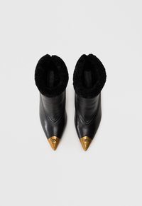 Botines de cuero negro hasta el tobillo con un puño negro peludo. Presentan una puntera puntiaguda con un acento dorado en la tapa de la puntera, y un detalle texturizado en el puño.
