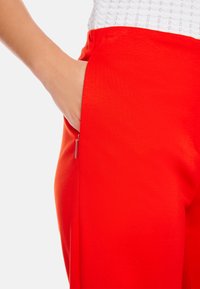 Rote Hosen mit glatter Textur, ausgestattet mit einer seitlichen Reißverschlusstasche. Der Bund sitzt hoch, und der Stoff wirkt leicht und atmungsaktiv.