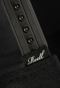 Schwarzer Stoffriemen mit strukturierter Oberfläche, der einen schwarzen Kunststoffabschnitt mit runden Nieten und einem weißen, bestickten "Reell"-Etikett aufweist.