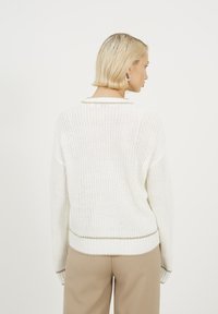 Biały sweter z dzianiny o luźnym kroju, z prążkowanymi mankietami i ozdobnym beżowym wykończeniem wokół dekoltu i dolnej krawędzi. Wzór o teksturowanym splocie.