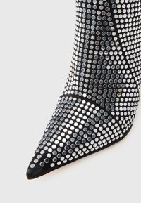 Pantof cu toc stiletto negru, cu vârf ascuțit, acoperit cu ținte argintii. Textură netedă cu captuseală din material textil negru și o siluetă elegantă.