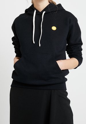 Mujer con sudadera negra con cordones blancos y un pequeño emblema circular amarillo en el pecho, con las manos en el bolsillo frontal.