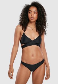 Črni bikini komplet, ki vključuje trikotno zgornji del z odrezanimi pasovi in ujemajoče se nizke hlačke. Gladek material z minimalnim okrasjem.
