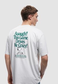 Camiseta de algodón blanca con texto verde en la parte posterior: "¡Aaaugh! ¡Este juego me vuelve loco!" con una ilustración de Snoopy.