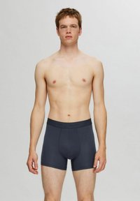 Tummanharmaat puuvillabokserit, joissa on joustava vyötärö, kuvattuna henkilöllä, jolla on neutraali ilme, seisomassa yksinkertaista taustaa vasten.