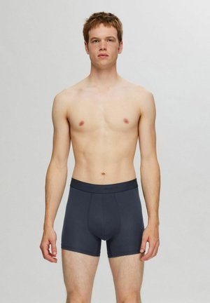 Boxers en coton gris foncé avec une taille élastique, présentés sur une personne avec une expression neutre, se tenant devant un fond uni.
