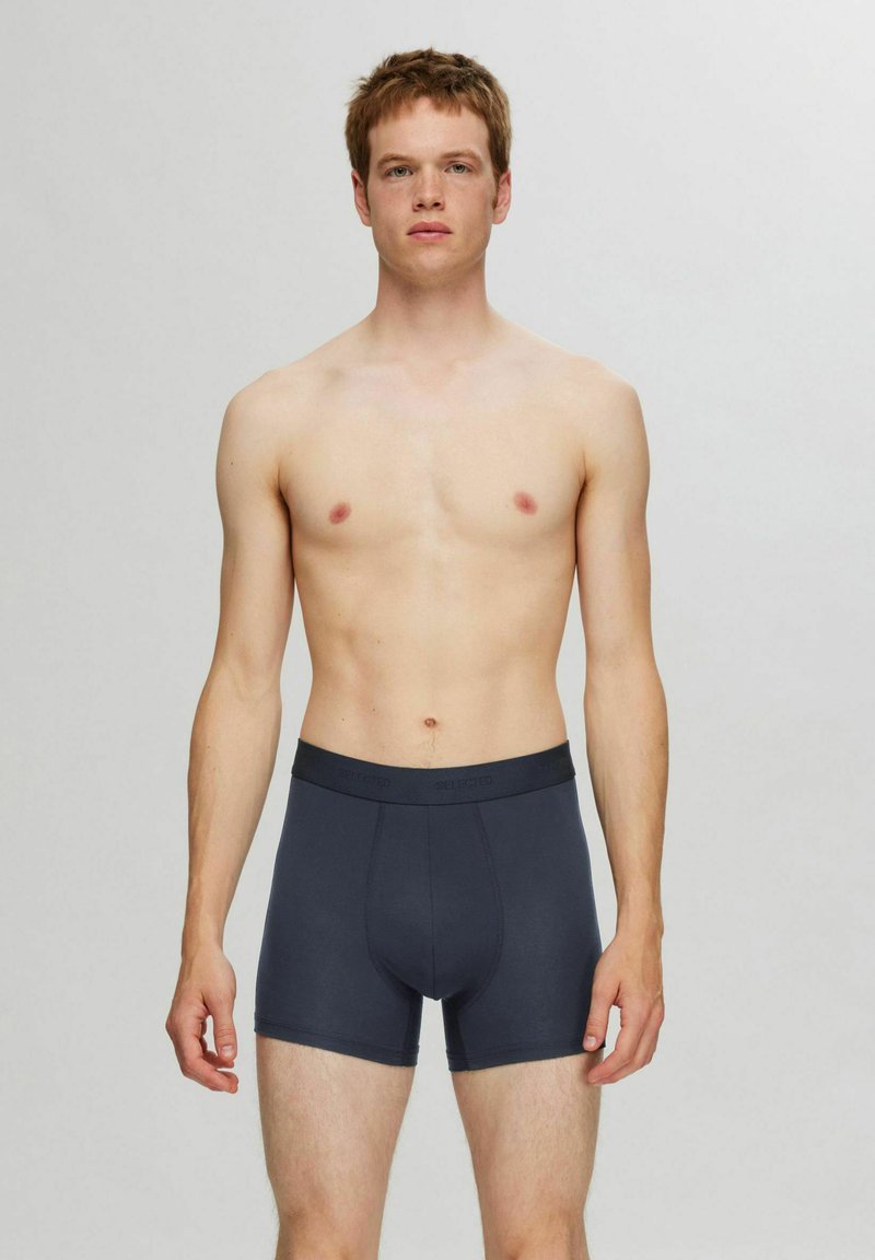 Tummanharmaat puuvillabokserit, joissa on joustava vyötärö, kuvattuna henkilöllä, jolla on neutraali ilme, seisomassa yksinkertaista taustaa vasten.