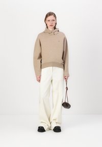 Beige hoodie met een kangoeroezak, gecombineerd met brede witte broeken en zwarte plateau schoenen. Model houdt een kleine bruine tas vast.