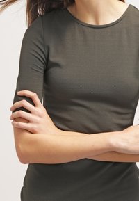 Ciemnozielona, fakturowana bluzka z krótkimi rękawami i okrągłym dekoltem, charakteryzująca się dopasowanym krojem. Ręce skrzyżowane z przodu.