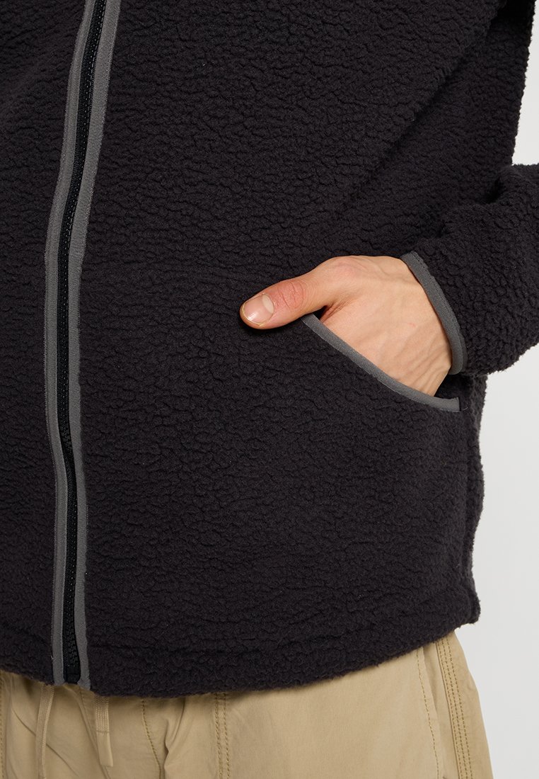 Giacca in pile nera con una superficie textureata, cerniera grigia e rifiniture grigie attorno alle tasche. Una mano è appoggiata in una tasca anteriore.