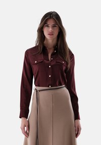 Woman with long brown hair wears a burgundy buttoned blouse and beige skirt with a thin black belt against a plain white background.