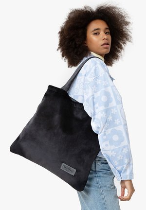 Bolso tote de ante negro con una textura suave, forma rectangular ancha y asas reforzadas. Presenta un parche con el logo en un lado.