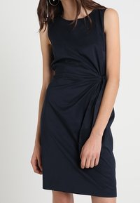 Granatowa sukienka bez rękawów o dopasowanym kroju, z drapowanym materiałem po jednej stronie i gładką strukturą. Minimalistyczny design, długość do kolan.