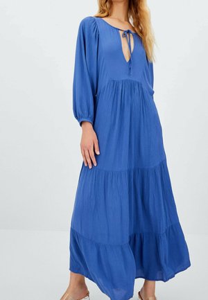 Vrouw draagt een lange, vloeiende blauwe jurk met lange mouwen en een strik bij de V-hals, staand tegen een effen lichte achtergrond.