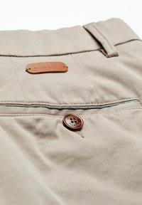Beiger Hosenprozess mit einem braunen Knopf und einem Lederetikett, auf dem der Markenname "bugatti" gestickt ist.