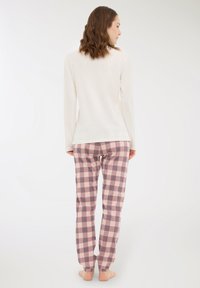 Donna con capelli castani mossi sta in piedi scalza, girata di spalle, indossando una maglietta bianca a maniche lunghe e pantaloni da pigiama a quadri rosa e grigi.