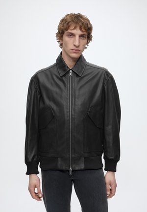 Junger Mann mit welligem hellbraunem Haar, der eine schwarze Lederjacke mit Reißverschluss und dunkle Jeans trägt, steht vor einem weißen Hintergrund.