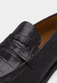 Mocassin en cuir noir à texture crocodile avec détail de sangle, montrant un gros plan de la partie supérieure et de la doublure intérieure.