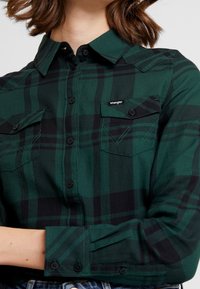 Camicia a quadri verde e nera in cotone, con chiusura a bottoni frontale, due tasche sul petto e dettagli cuciti sulle spalle e sulle maniche.