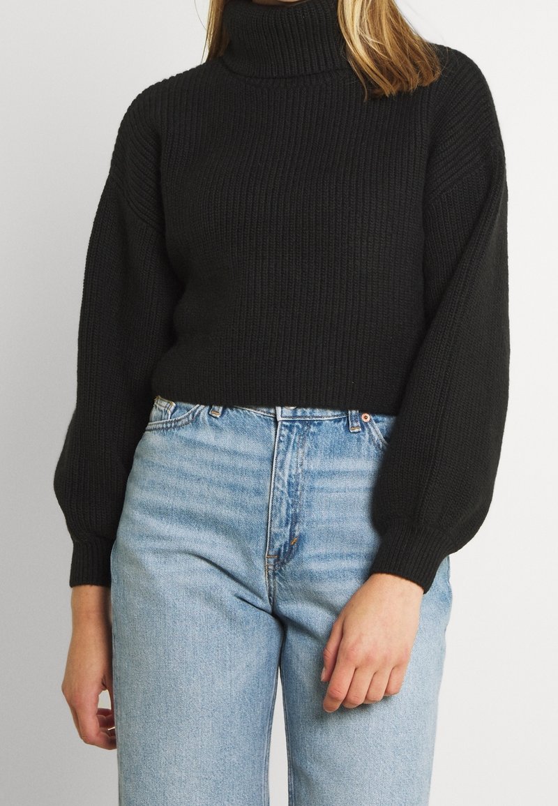 Femme portant un pull à col roulé noir côtelé avec des manches ballon et un jean taille haute bleu clair sur un fond uni.