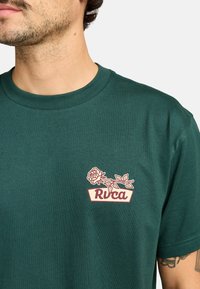 Camiseta de algodón verde con un gráfico de rosa roja y blanca y el texto "Rvca" en el lado izquierdo del pecho. Diseño de manga corta y cuello redondo.