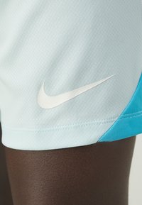 Primer plano de un muslo de piel oscura que lleva pantalones cortos deportivos Nike de color azul claro con el logotipo blanco del swoosh de Nike y ribetes azules.