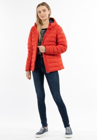 Rød pufferjakke med hætte og quiltet design, med en frontlynlås, iført mørke jeans og grå sneakers.