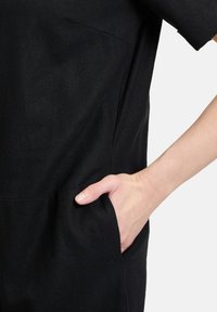 Czarna tkaninowa odzież o luźnym kroju, krótkich rękawach i kieszeni bocznej. Tekstura wydaje się gładka, a design jest minimalistyczny.