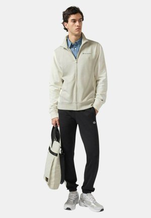 Felpa beige chiaro con zip, pantaloni neri scuri, porta una borsa chiara. Sneakers con accenti grigi e bianchi. Design semplice, branding minimale.