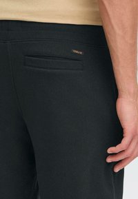 Pantalones de chándal negros con una textura suave, cinturilla elástica y un bolsillo trasero. Una pequeña etiqueta marrón con la palabra "SOLID" es visible en la cinturilla.