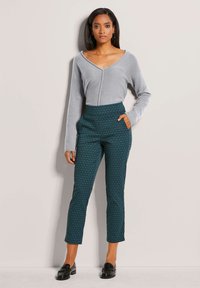 Graues Langarmoberteil mit V-Ausschnitt, kombiniert mit tealfarbenen gemusterten Hosen mit seitlichen Taschen. Schwarze Slipper runden den Look ab.