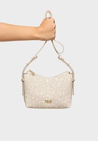 Bolso de mano de cuero beige con patrón embosado, detalles en hardware dorado y cierre con cremallera. Una mano sostiene la correa para referencia de tamaño.