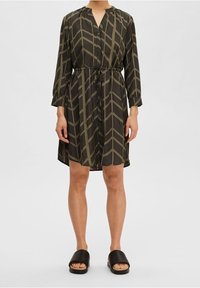 Abito verde a fantasia con maniche lunghe, scollatura a V, vita con cordino e una silhouette ampia, abbinato a sandali neri slip-on.