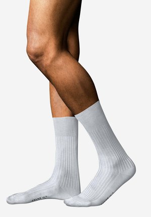 Persona con calzini bianchi a coste al ginocchio, in piedi con gambe incrociate su uno sfondo bianco uniforme.