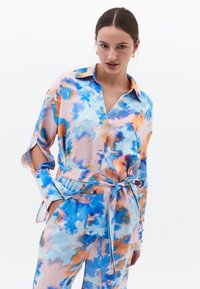 Kleurrijke outfit met strikceintuur in een abstract patroon van blauw, oranje en roze. Heeft lange mouwen met inkepingen en knoopdetails. Zijden textuur.