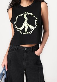Sort cropped tank top med et grønt grafisk design af et fredssymbol omgivet af kæder. Kombineret med højtaljede sorte denimbukser.