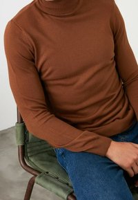 Maglione a collo alto marrone con texture a coste, abbinato a jeans blu. Il maglione ha maniche lunghe e un design aderente.