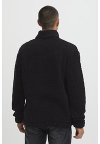 Pullover in pile nero con colletto alto, superficie testurizzata e maniche lunghe. La parte posteriore è piatta senza ulteriori dettagli.