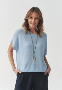 Helles blaues gestricktes Sweater mit kurzen Ärmeln, lockerer Passform und geripptem Halsausschnitt, getragen über dunkelblauen Hosen; dazu ein Anhänger-Halsband.