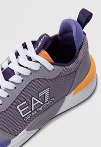 Vijolični športni čevelj z mrežnim zgornjim delom, belimi vezalkami, oranžnimi poudarki in gumijastim podplatom. Na njem je umesčen logo EA7 Emporio Armani.