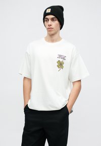 Biała bawełniana koszulka z krótkim rękawem, z zielonym motywem czterolistnej koniczyny i czarnym napisem na lewej piersi. Czarna czapka beanie założona na głowę.