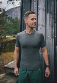 Hombre vestido con una camiseta gris y pantalones cortos verdes se encuentra al aire libre junto a una pared de madera, mirando a su derecha con una ligera sonrisa.