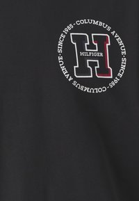 T-shirt nera con un logo rotondo che presenta una "H" bianca e accenti rossi, bianchi e blu, testo: "HILFIGER COLUMBUS AVENUE DAL 1985."