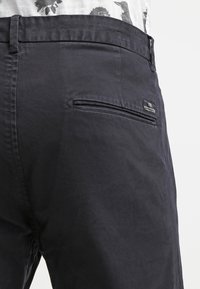 Dark navy pants made of a smooth, textured fabric. Features a zippered back pocket and a small branded label near the pocket.