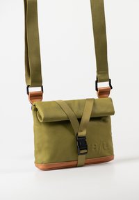Olivgrüne Tasche mit einem Rolltop-Design, braunem Lederboden, verstellbarem Schultergurt, schwarzem Schnallenverschluss und geprägtem Logo auf der Vorderseite.