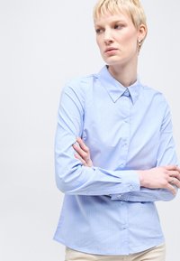 Γυναίκα με κοντά ξανθά μαλλιά, φορώντας ένα γαλάζιο πουκάμισο με ρίγες και κουμπιά, στέκεται με σταυρωμένα τα χέρια, κοιτάζοντας προς τα πλάγια πάνω σε απλό φόντο.