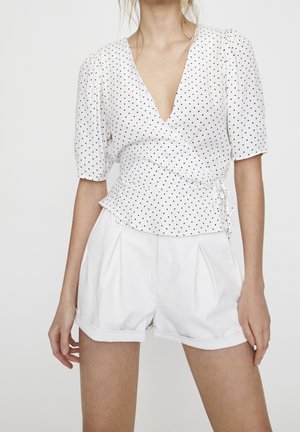 Vrouw draagt een witte wikkelblouse met zwarte polkadots en hoge, geplooide witte shorts.