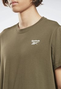 Osoba nosící olivově zelené tričko Reebok s bílým logem na hrudi, zobrazená od krku po horní část trupu na jednobarevném pozadí.